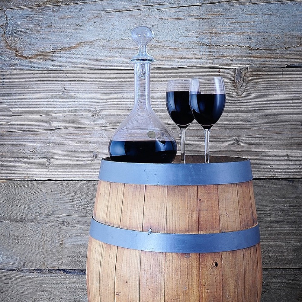 Un decantador de cristal con vino tinto y dos copas sobre un barril de madera rústico.