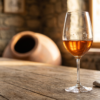 Copa de vino naranja (orange wine) sobre una mesa rústica de madera con vasija tradicional de arcilla al fondo.