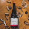 Botella de vino sobre una mesa de madera rodeada de varios tipos de corcho, un sacacorchos y copas vacías.